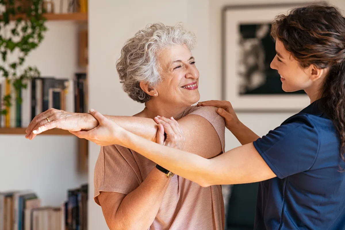 Happy senior woman doing exercise at home with physiotherapist. Old retired lady doing stretching arms at home with the help of a personal trainer during a rehabilitation session.